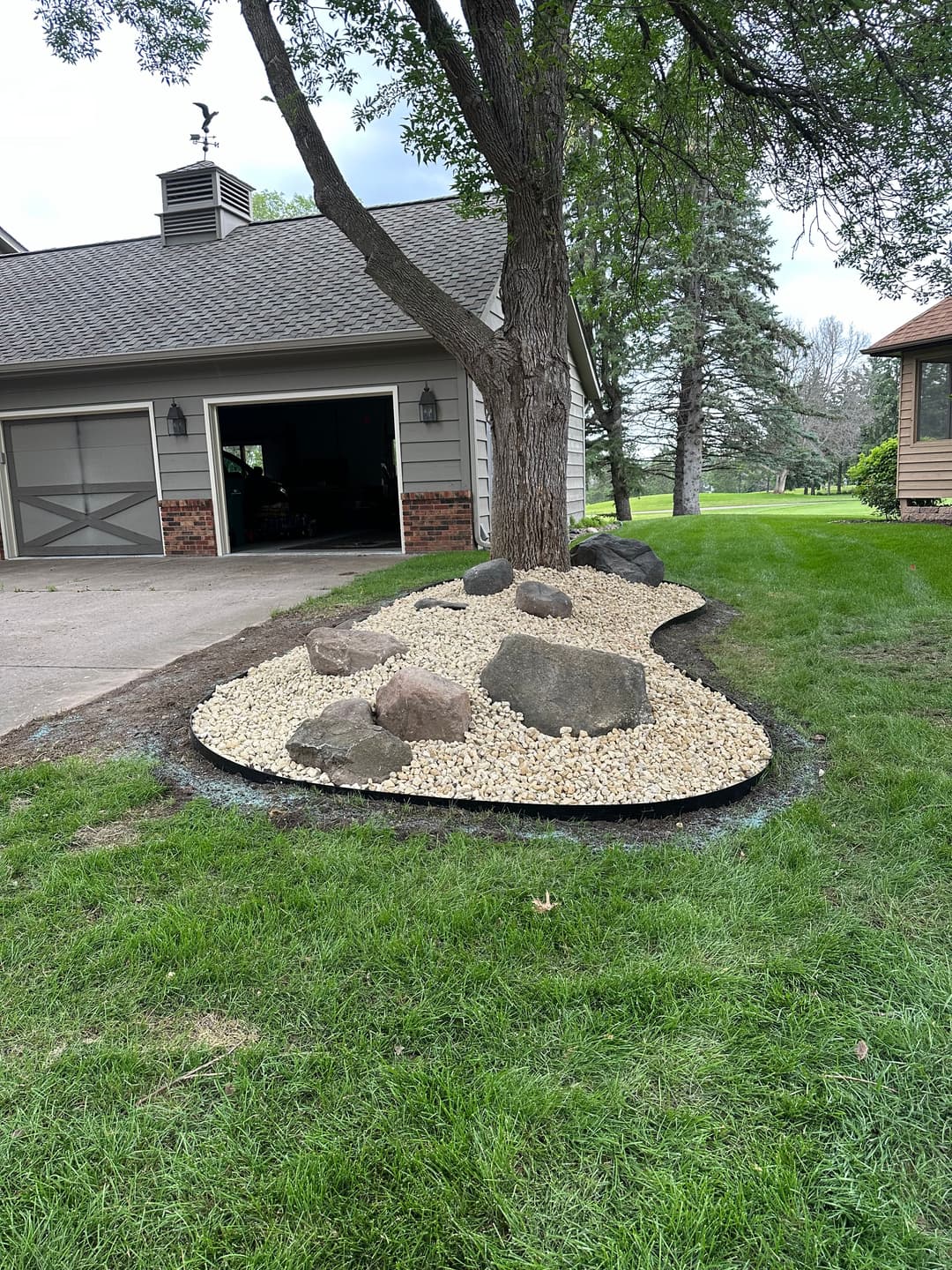 Landscaped garden with gravel, rocks, and a tree near a house and driveway.