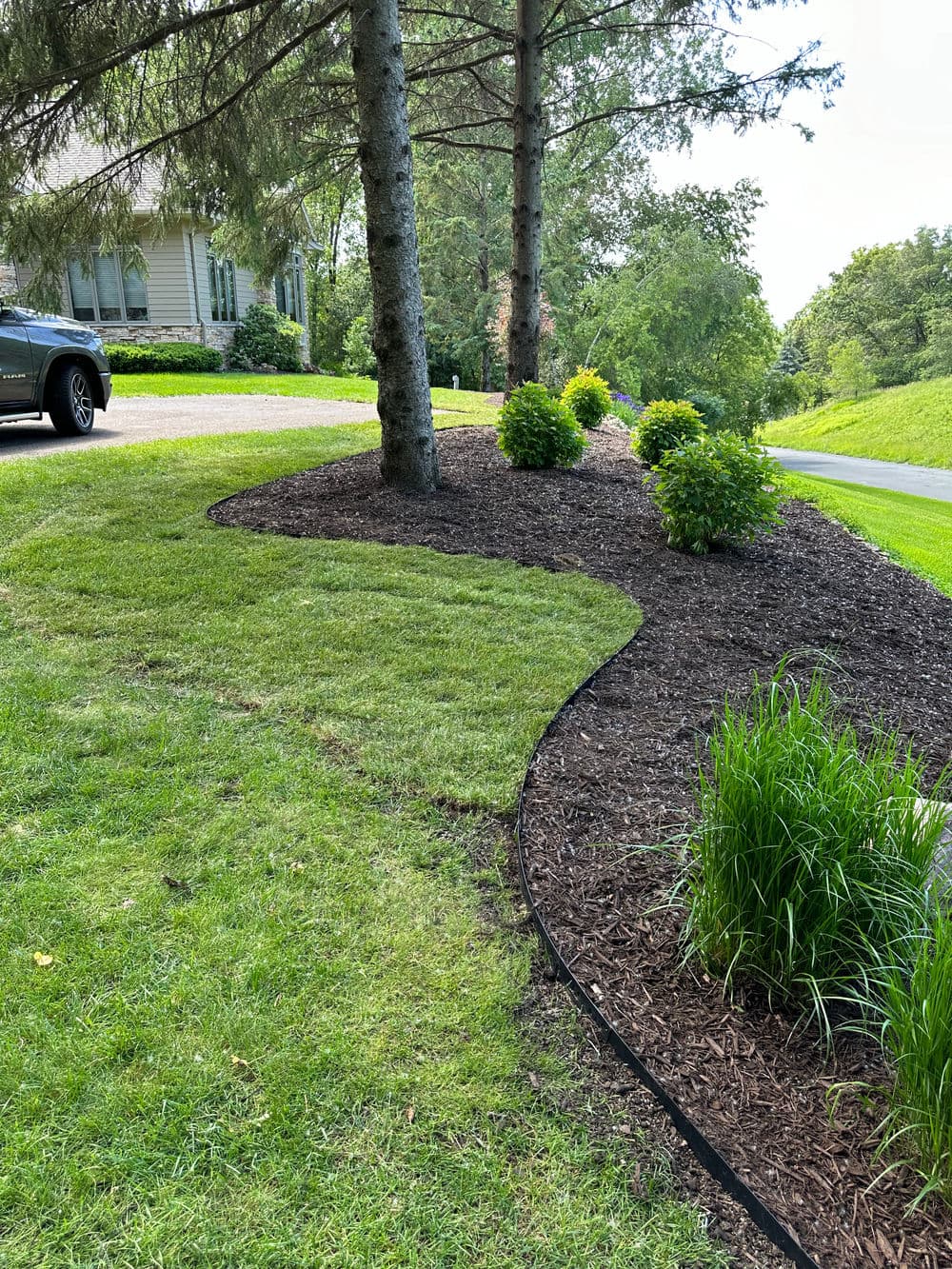 Curved landscaping with mulch, bushes, and grass along a driveway in a sunny area.